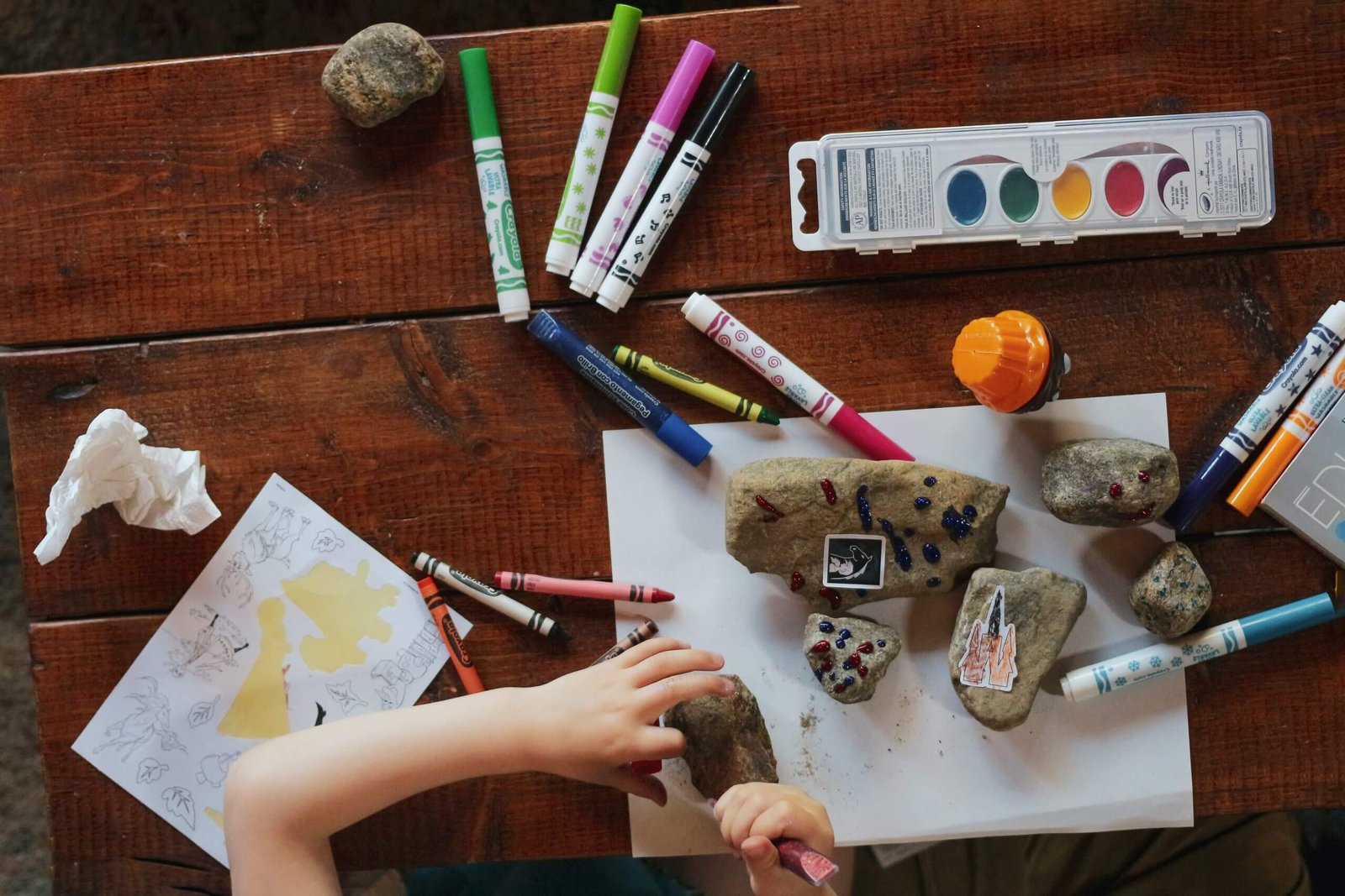 table with art supplies on it, child's hands painting rocks