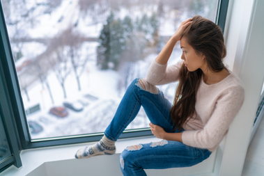 Person in long-sleeved tee, ripped jeans, and socks sitting in a window sadly looking down at everything covered in snow.